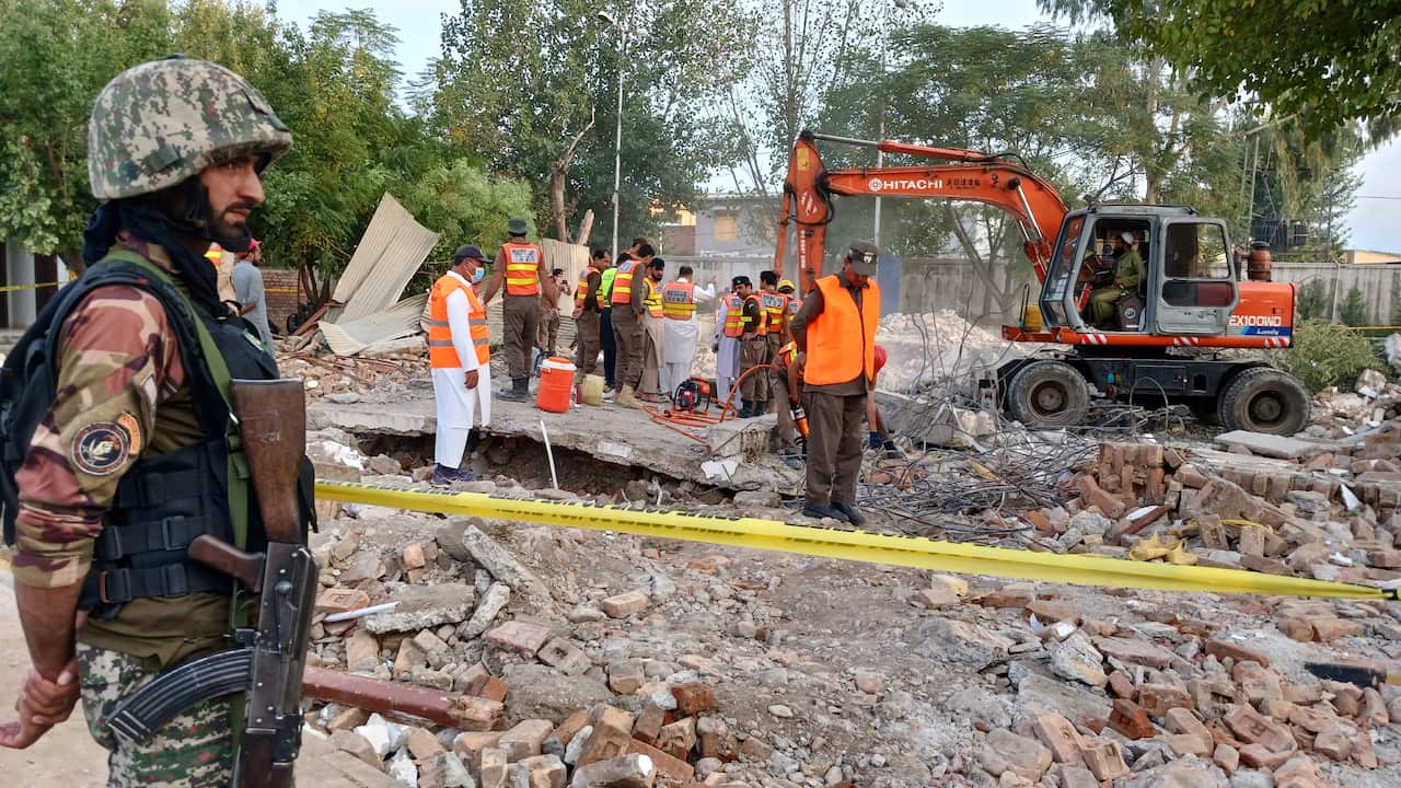 A person in military uniform looks on as rescuers work amid the debris of a destroyed building.