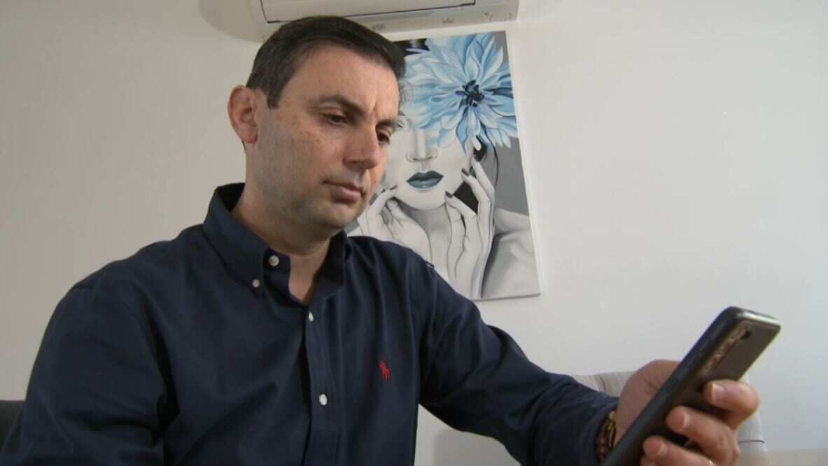 A man in a dark blue collared shirt looks down at a smartphone in his left hand.