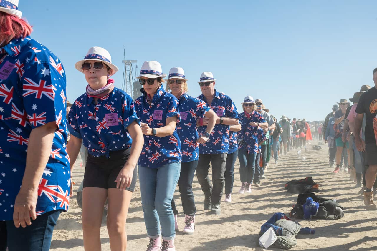 Lines of people, many wearing shirts featuring the Australian flag, doing the nutbush on dusty ground.
