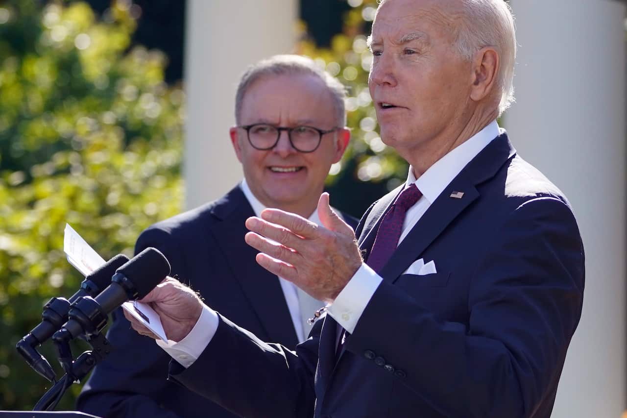A close-up photo shows two men in dark suits standing at a podium in daylight.