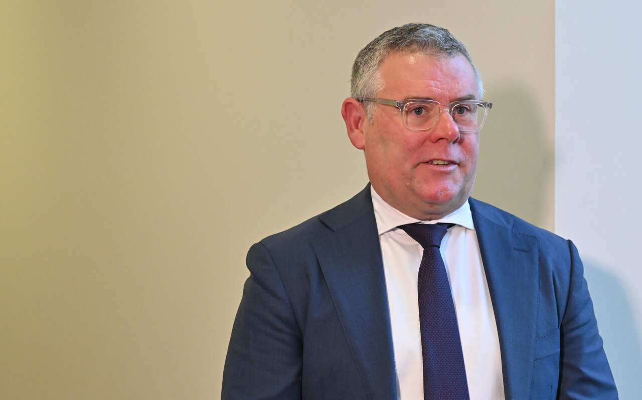 A person in a blue blazer and tie and white shirt standing in front of a light yellow background.