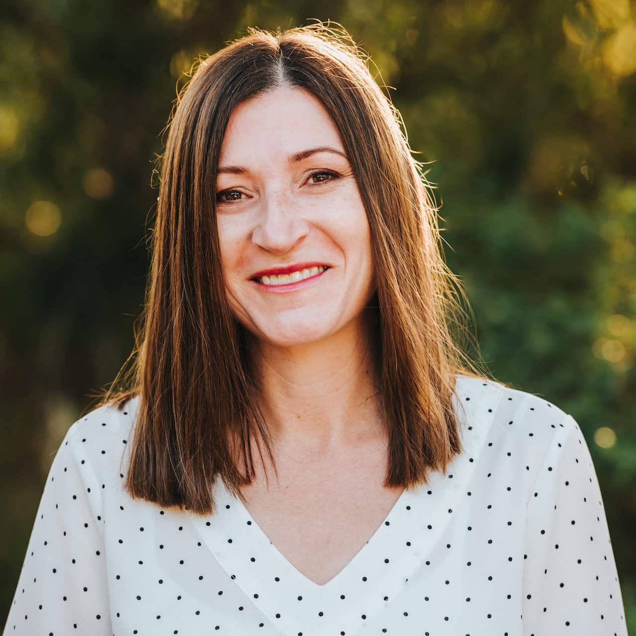 A woman with shoulder-length straight brown hair wearing a white polka-dot shirt.