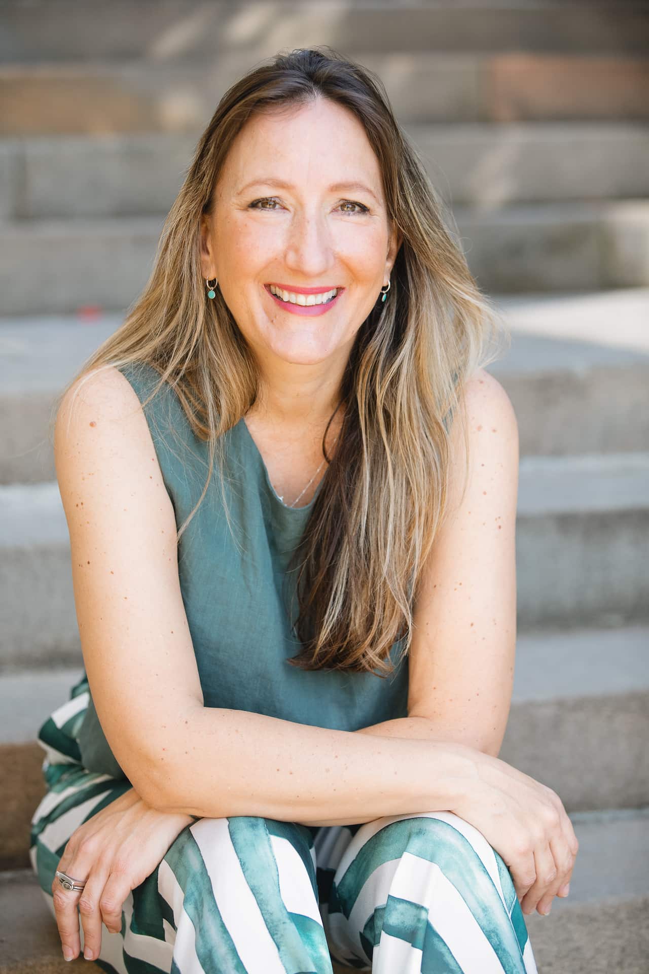 A smiling woman, sitting on stairs with her arms sat on her knees.