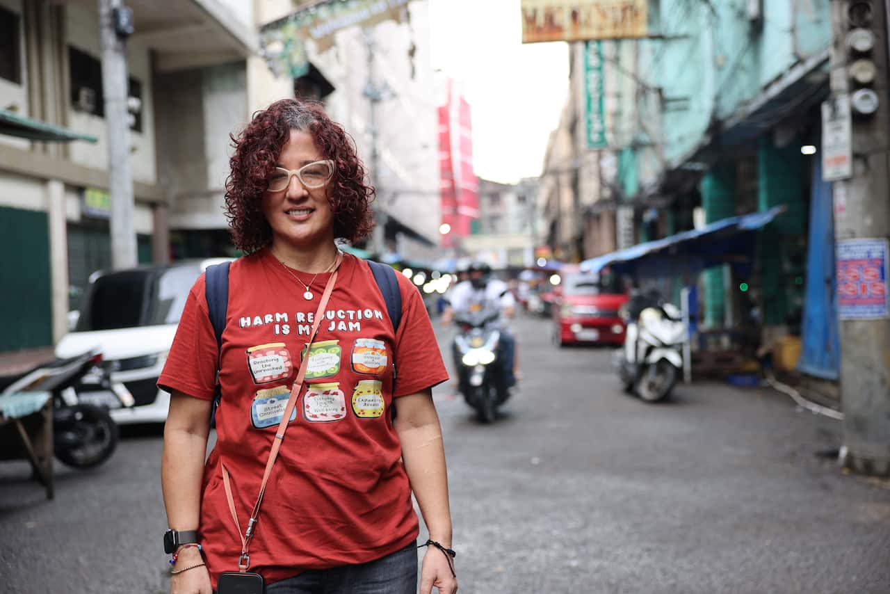A woman with curly hair coloured dark red stands in the middle of a street. A man on a motorbike is behind her. She is smiling, and wears large white glasses and a red t-shirt with pictures of jam jars on it. The t-shirt has text readinh 'harm reduction is my jam'. 