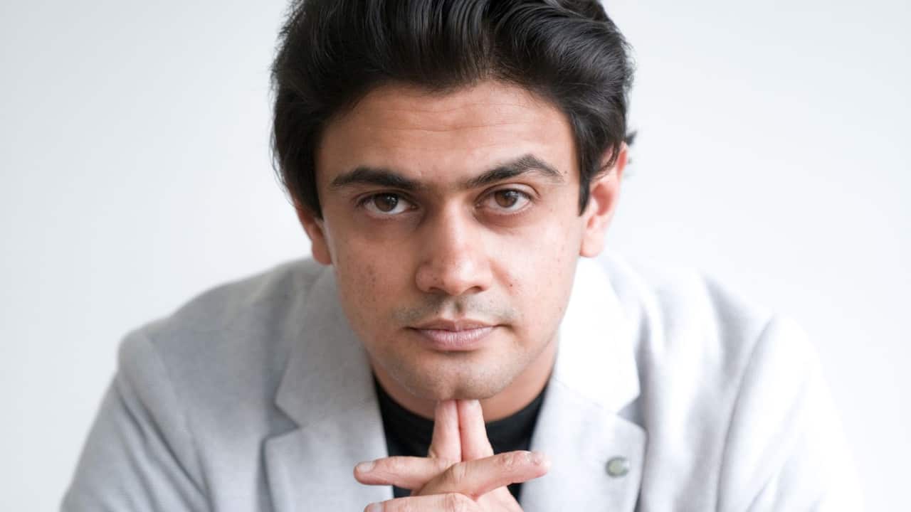 close up headshot of man in a grey blazer looking into camera, resting his chin on his crossed palms