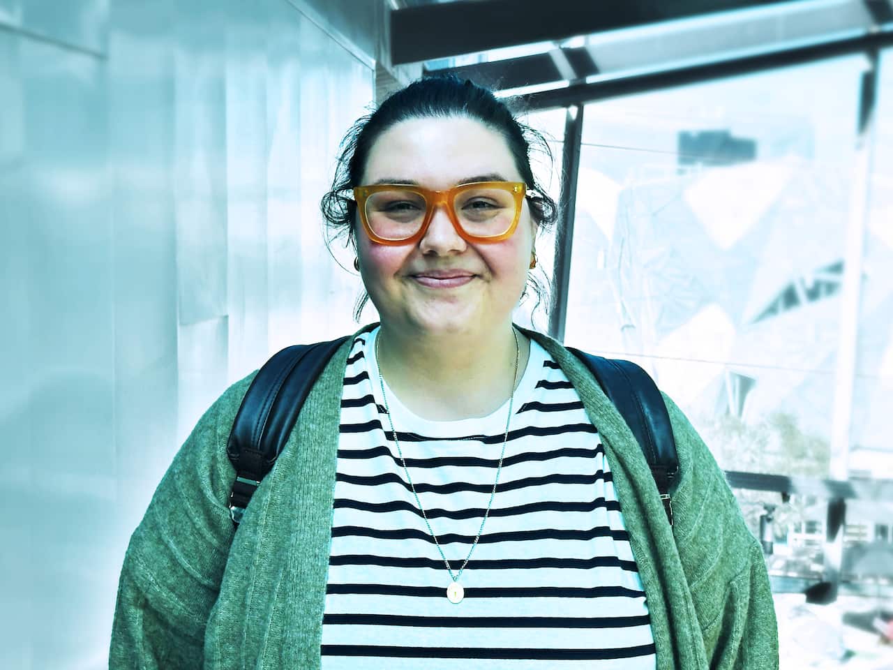 A person wearing a green cardigan, black and white striped top and amber-coloured glasses is smiling.