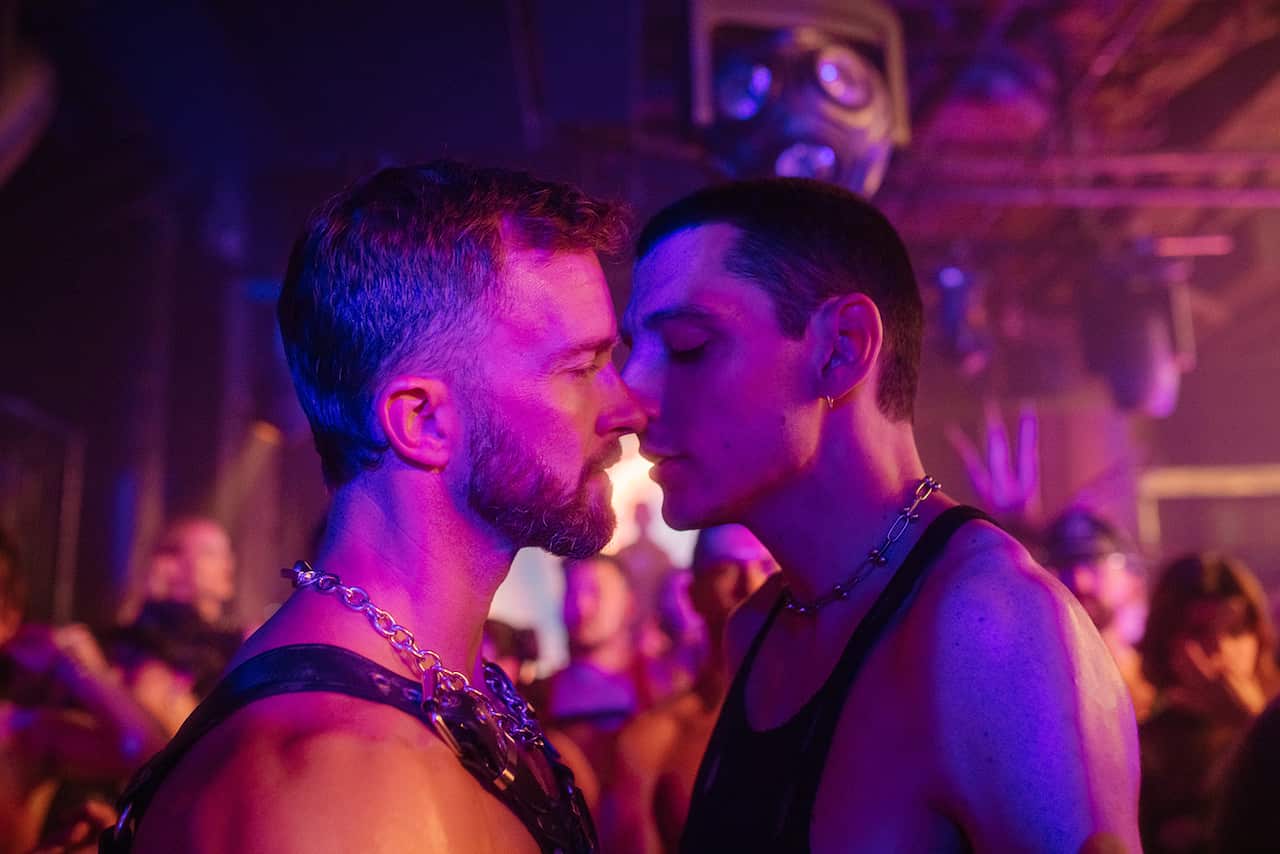 Two men close, face-to-face, stand in a room. The lighting is dark, with pink and purple tones.  