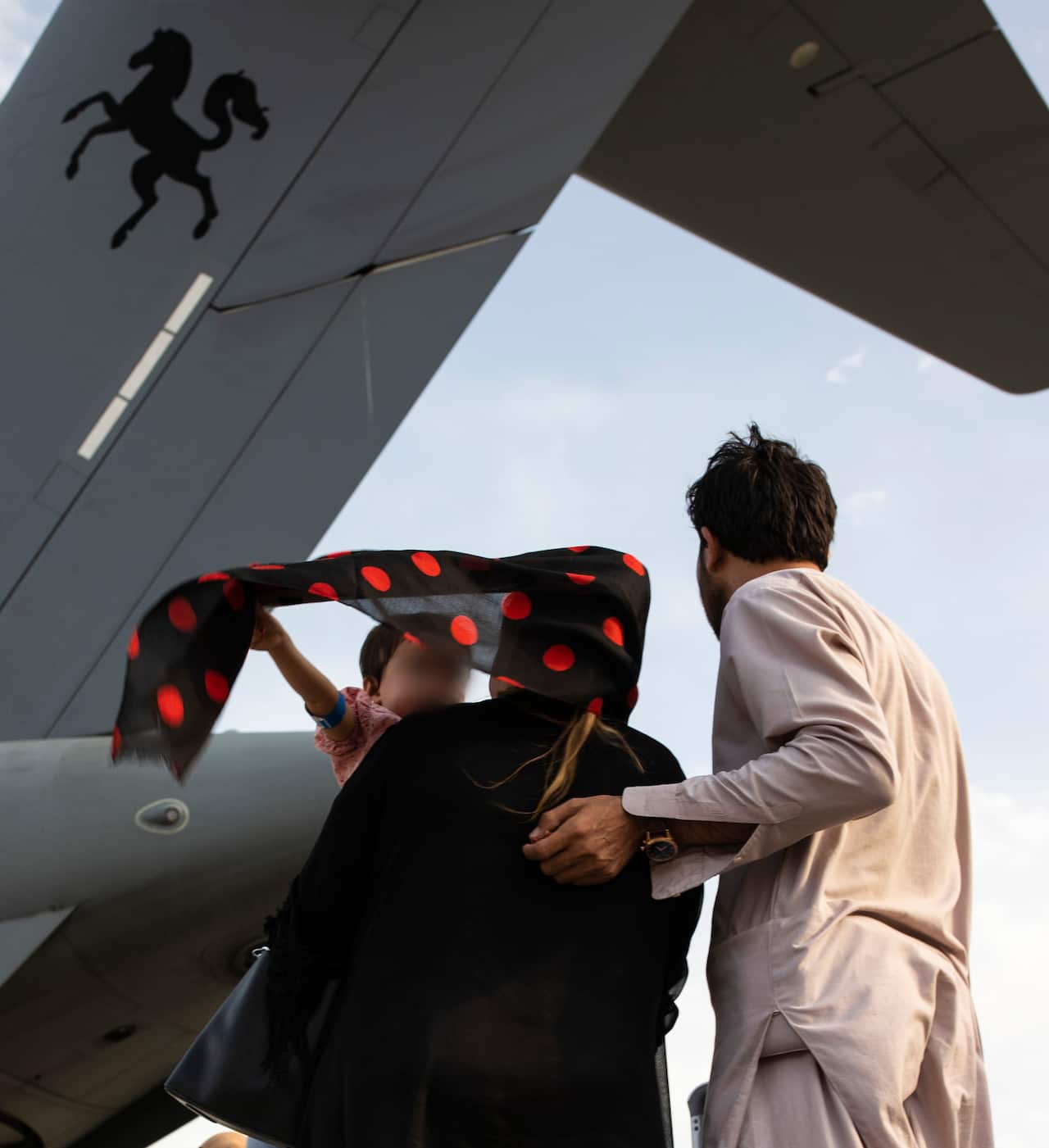 Child holds a scarf in his mother's hair as the father embraces her under a plane wing.