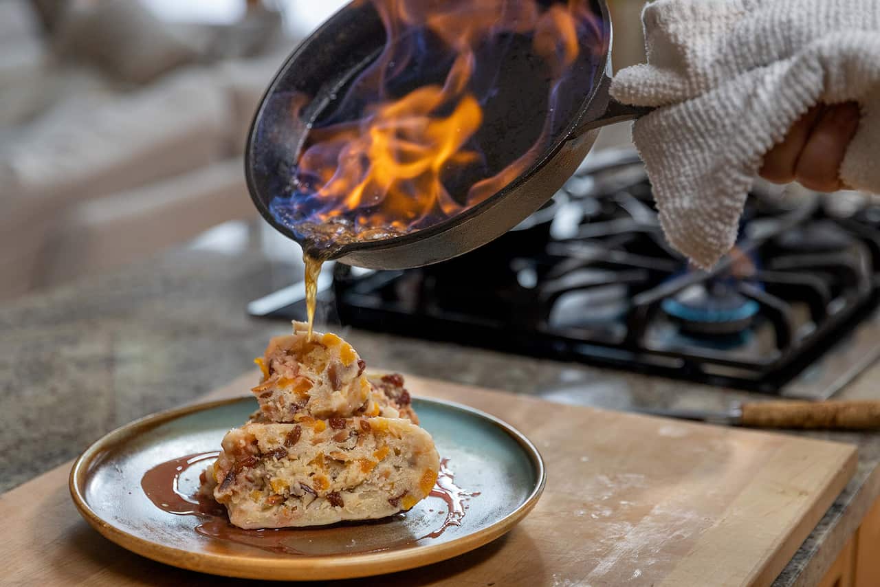 A slice of fruit-studded pudding sits on a plate. A saucepan with a flaming sauce is tilted over the plate. 