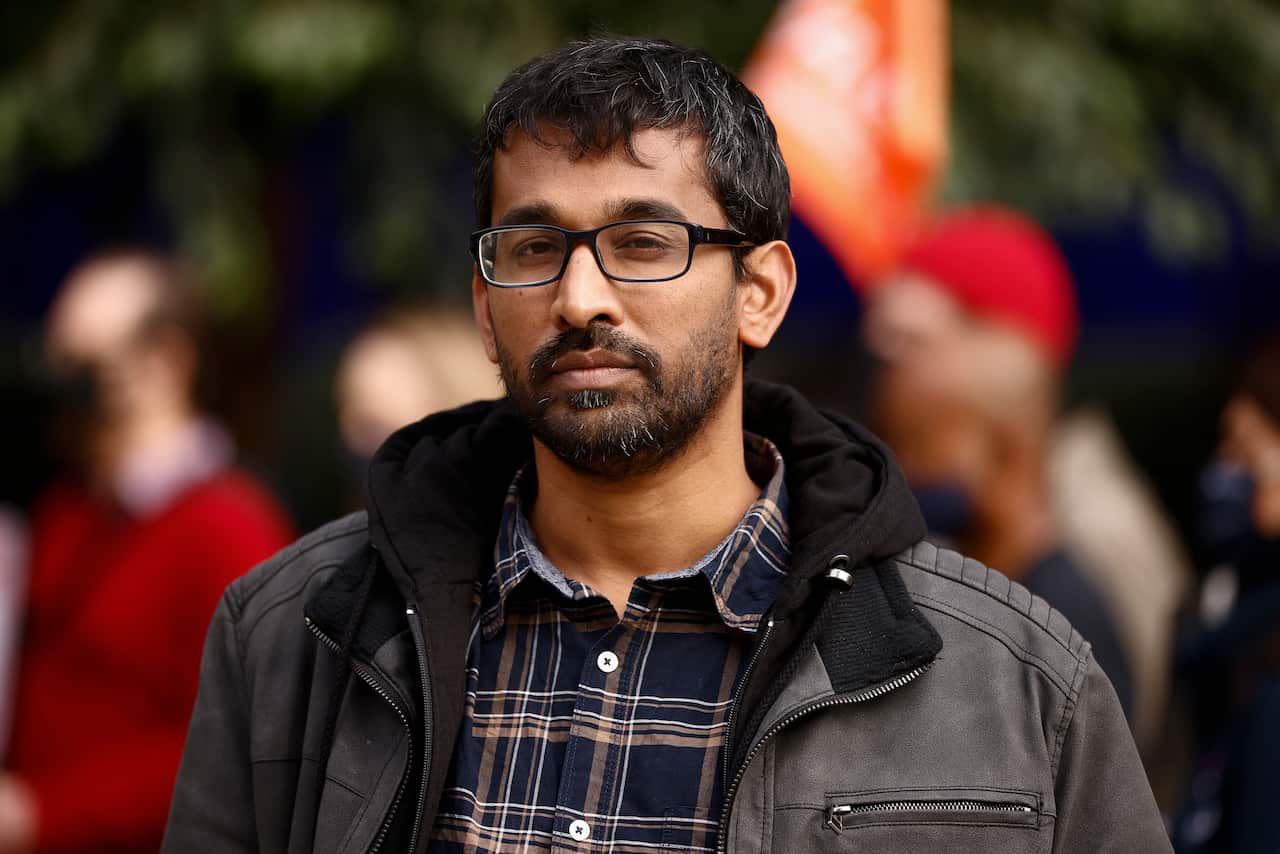 Aran gives a serious facial expression, wearing a jacket and tartan shirt.