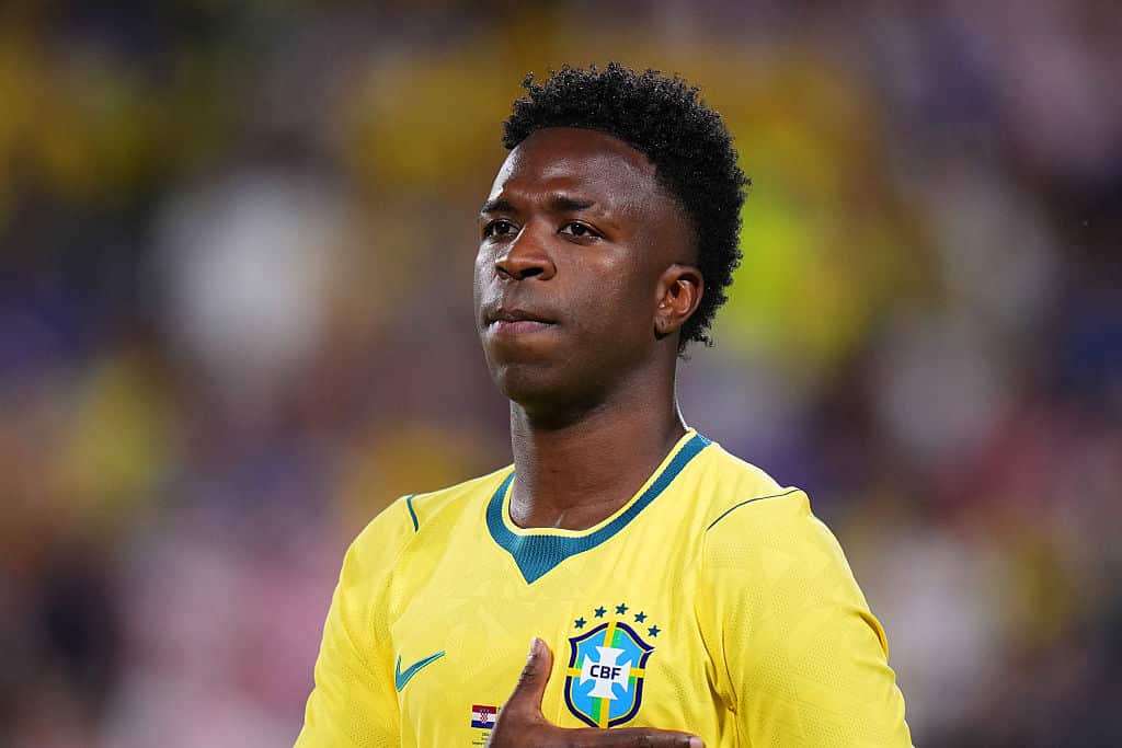 A footballer wearing a yellow jersey with green collar and holding his right hand on his left chest 