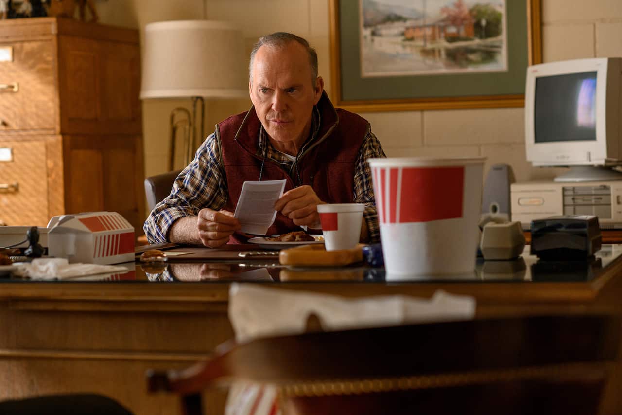 A man in a check shirt and red vest sits at a desk; the older-style computer beside him indicates this is set in the past. 