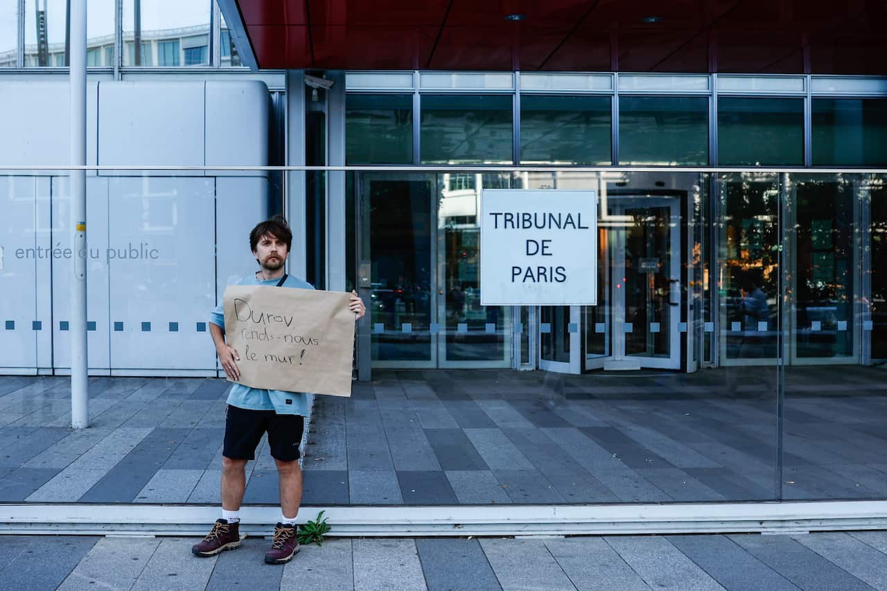 Man holding sign in French stands in front of a glass building and another sign that reads 'Tribunal De Paris'