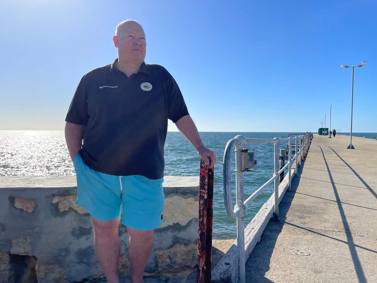 Middle-aged white man standing on a jetty with hand on railing. He is wearing a dark blue polo shirt and light blue shorts