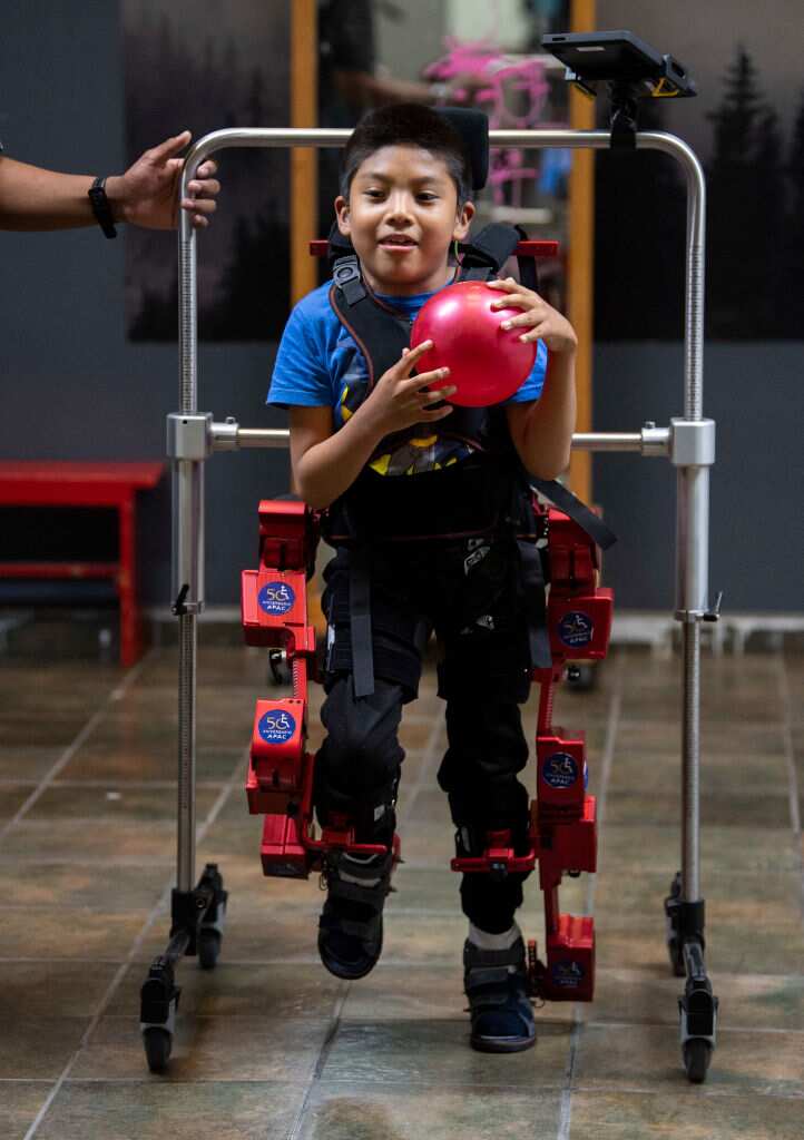 An eight-year-old boy with cerebral palsy is using a robotic frame that helps him to walk. An adult is holding the frame. 