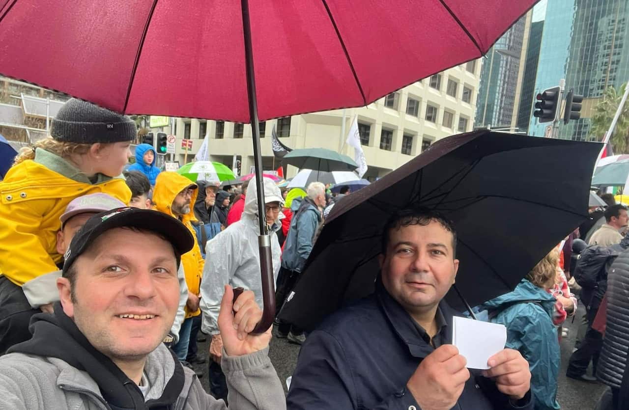 Two men taking a selfie under umbrellas while participating in a rainy street protest, with a crowd and city buildings in the background.