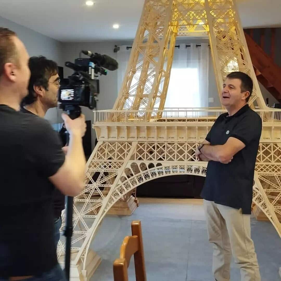 A man being interviewed for tv, standing in front of the eiffel tower he constructed out of matchsticks.