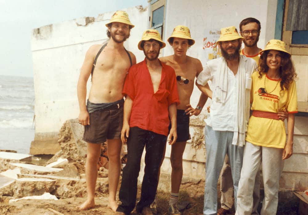 Five men and a woman standing in front of a dilapidated building by the ocean.