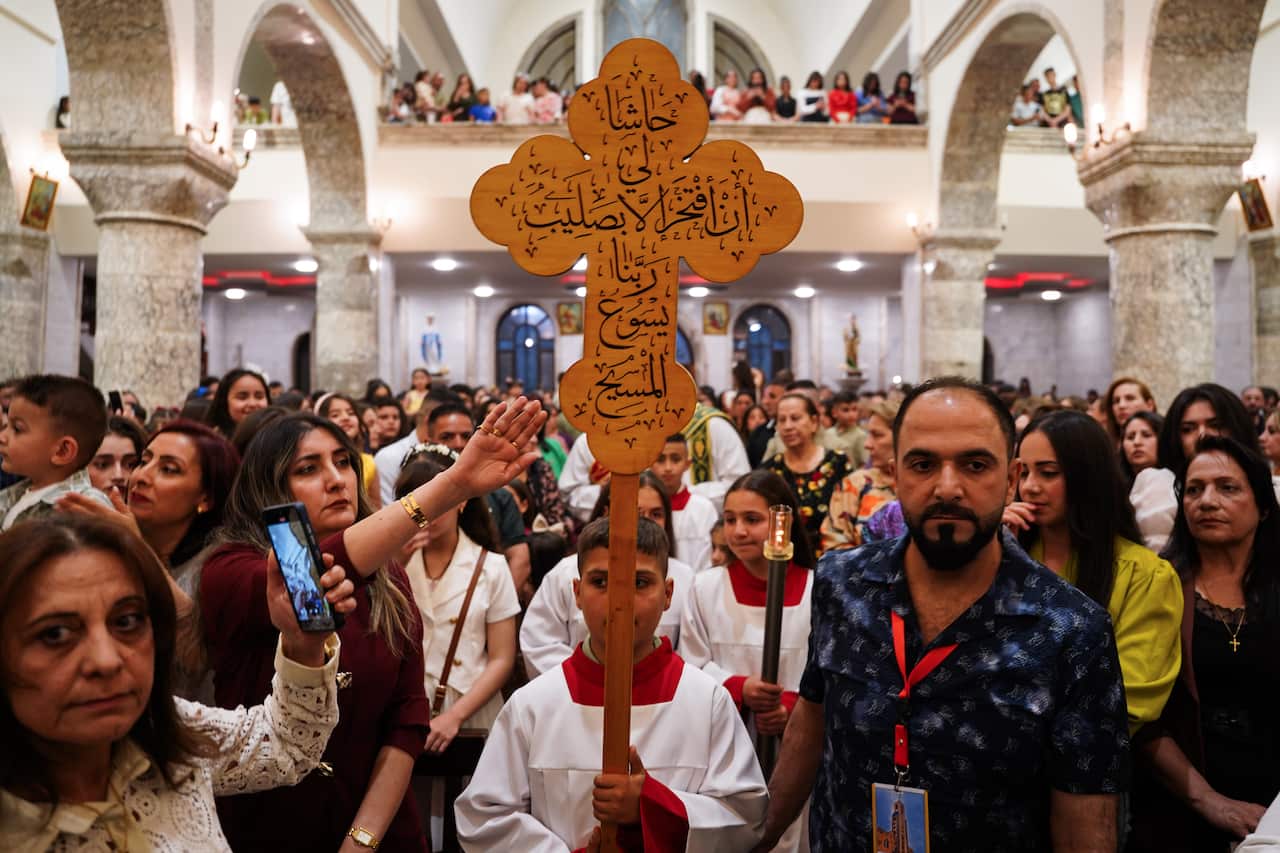 A young altar boy in a white robe standing in a crowd of Christian churchgoers, holds up a crucifix with Arabic writing. 