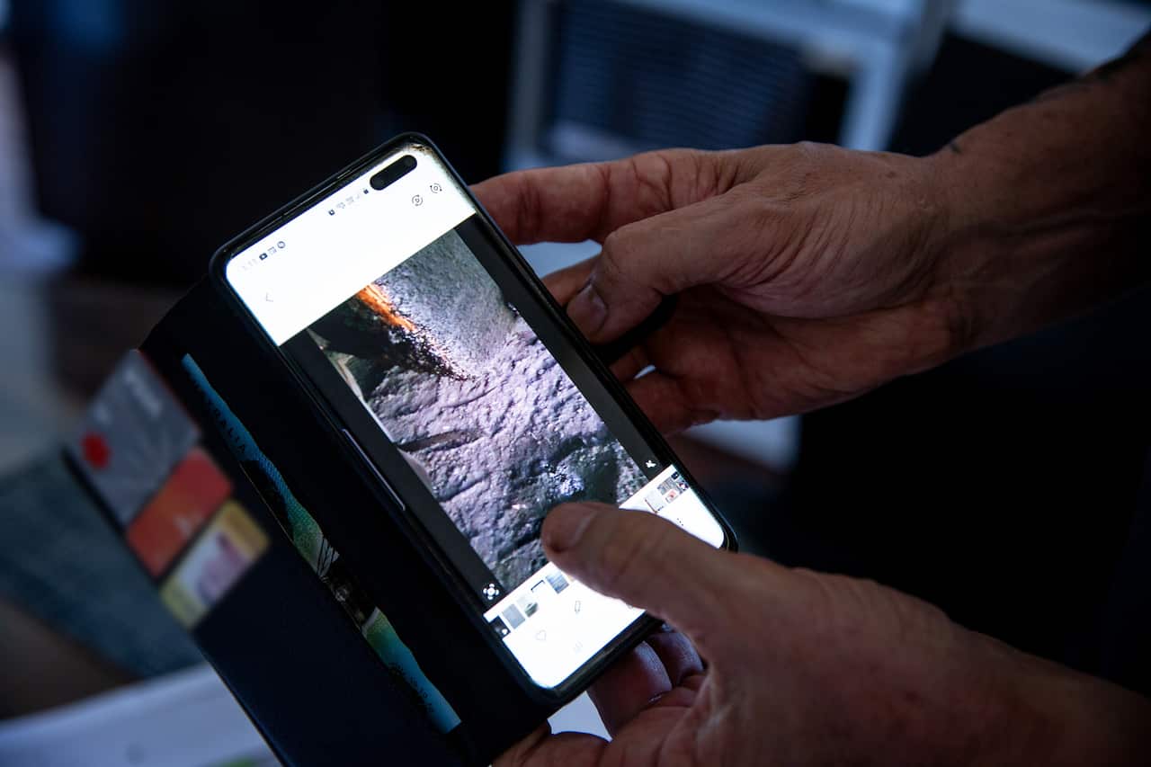 A close-up of a man holding a phone with its screen displaying a photo of the mechanic's workshop at a coal mine filled with dust.