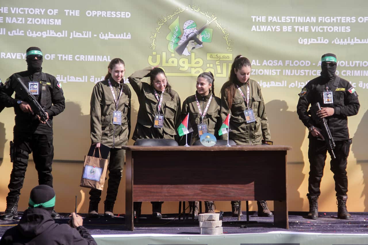 Four women wearing green military fatigues stand on a stage flanked by two men wearing black balaclavas, green headbands and carrying rifles. 