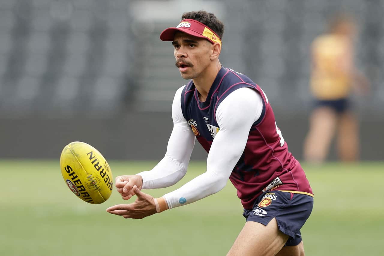 Man in maroon and navy blue Brisbane Lions jersey and matching cap throws football. 
