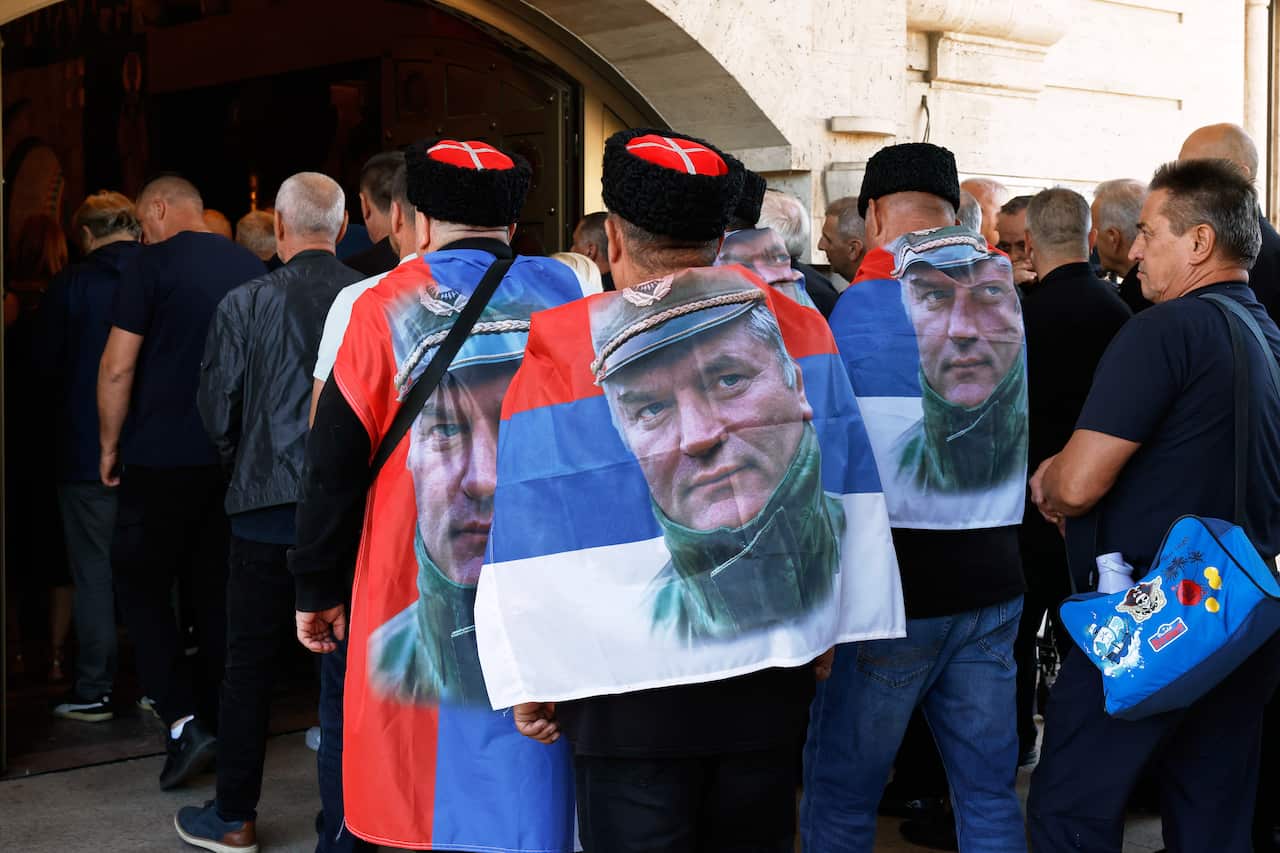 A medium shot capturing a crowd of people from behind gathered outside an arched doorway. In the foreground, three men stand facing away from the camera, wearing black fur style traditional caps (shubara)—two featuring red tops with a white cross design. Draped across their backs like capes are red, blue, and white tricolor flags, each printed with a large, prominent face of a man in a military cap and jacket. To the right, another man stands in profile looking toward the left.