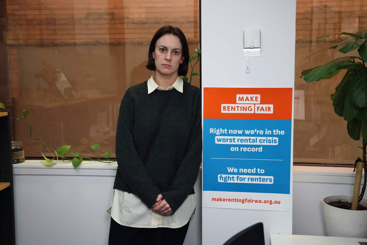 A lady with a sweater and collared inner shirt, leans against a wall with a prominent poster.
