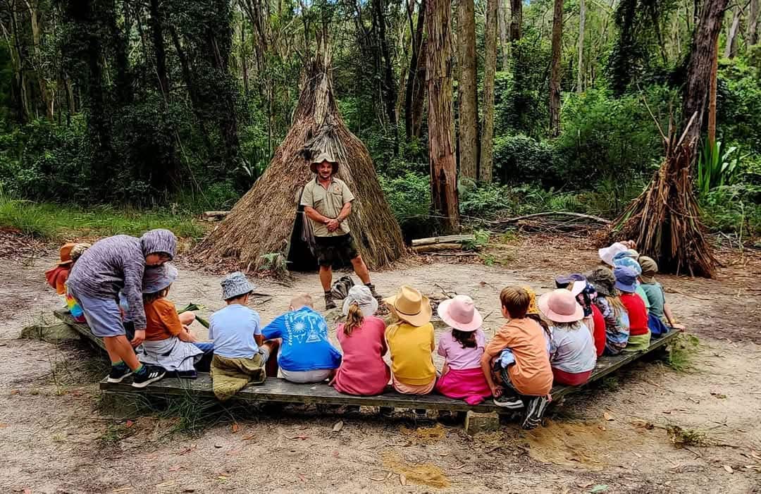 A group of kids siting in a circle in the bush while a man speaks to them.