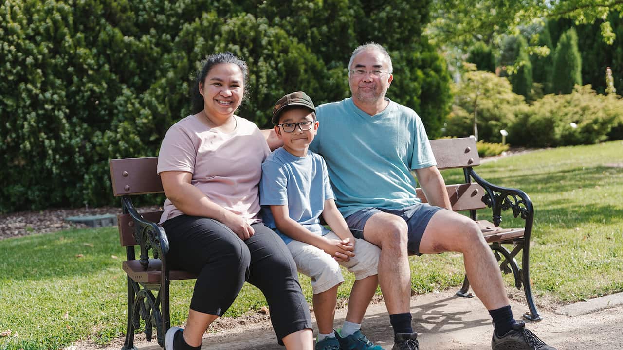 A smiling mand and woman sit on a park bench, with a boy sitting between them. 