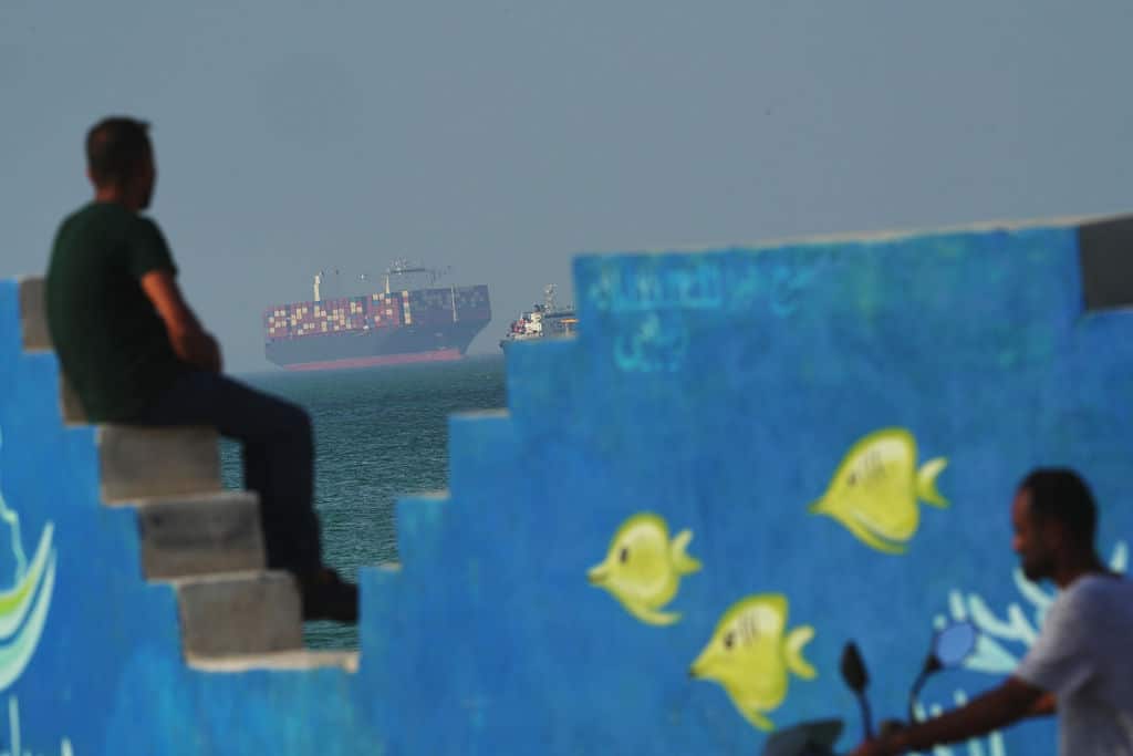 A man behind a mural depicting fishes at the shoreline watches as a cargo boat navigates the sea.
