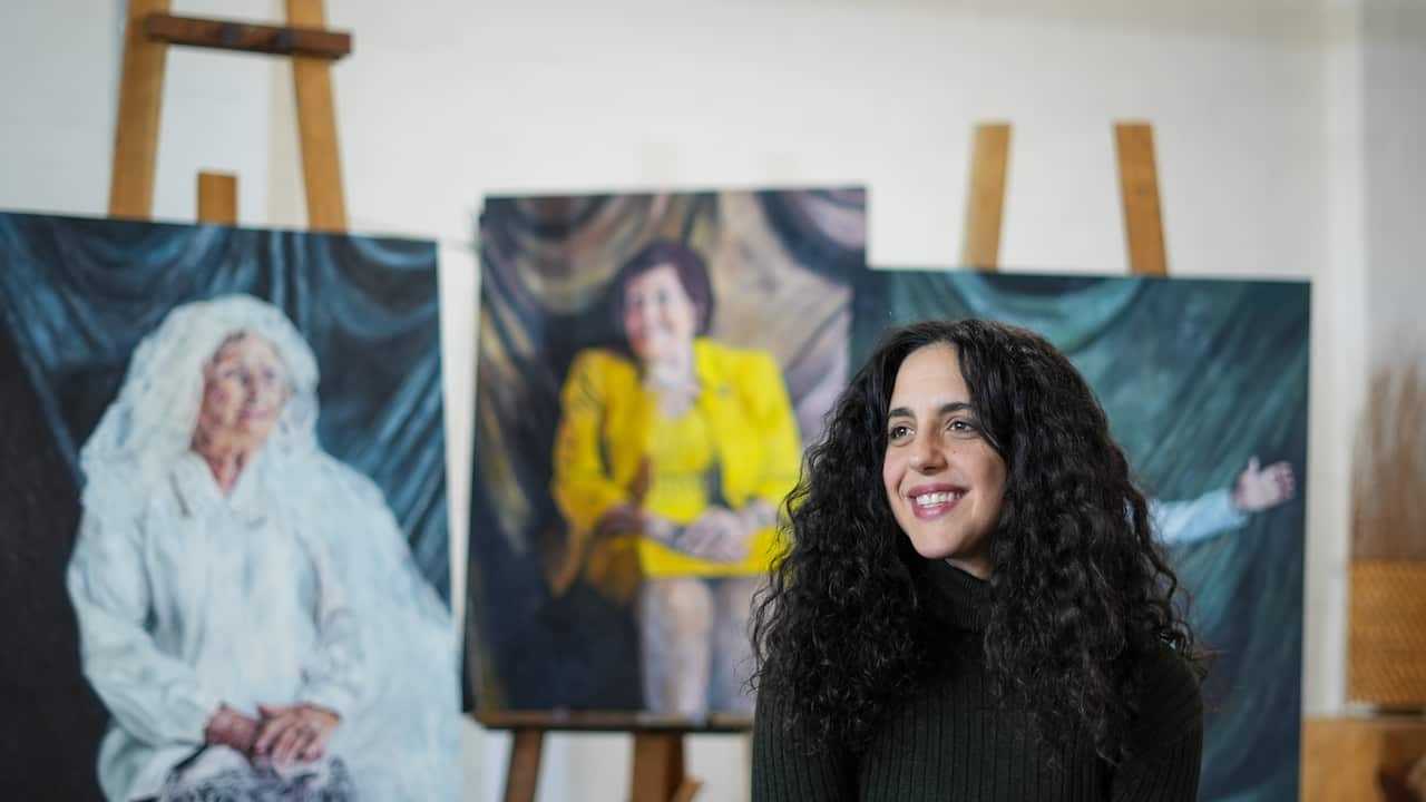 A woman with long, dark, curly hair smiles. Behind her are three portraits on wooden easels