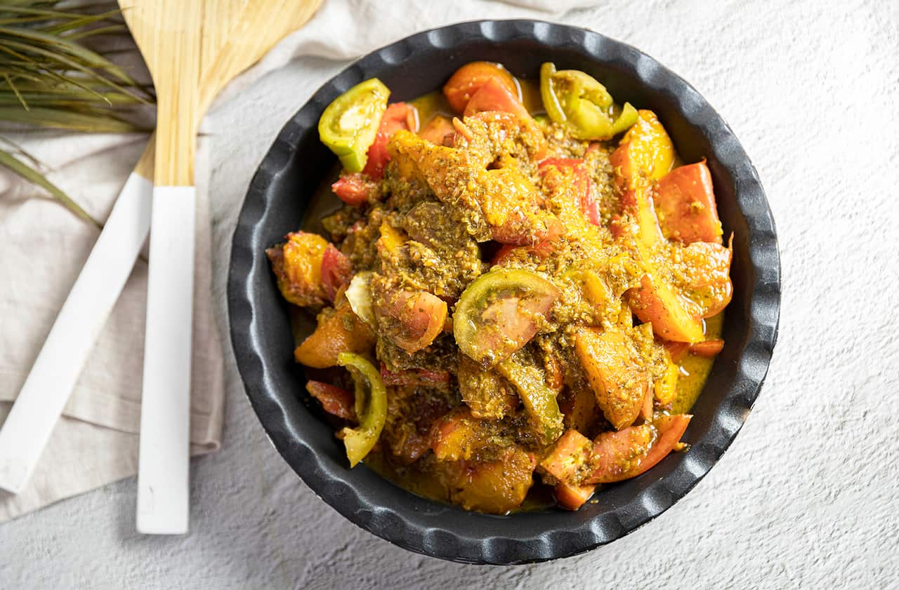 A tomato and peach salad, with a thick sauce, sits in a dark bowl on a white surface. Salad servers sit alongside, on a napkin. 
