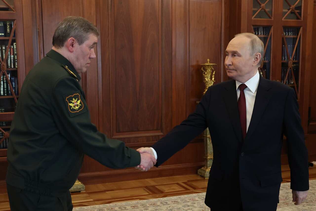 A man in a black suit (right) shaking the hands of a man in army greens, in an office with wooden walls.