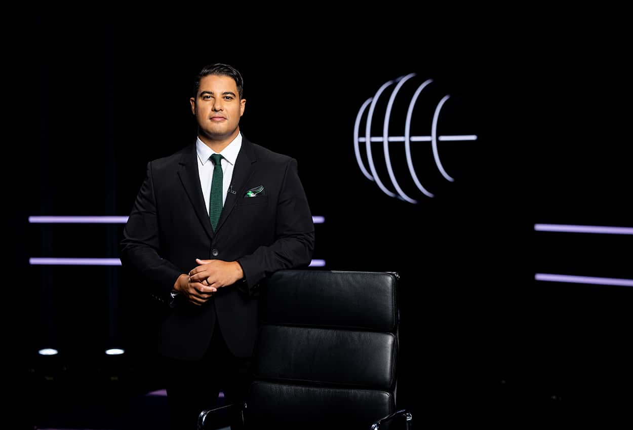 Marc Fennell, in a dark suit with a white shirt and green tie, stands on a dark set, beside a black chair.