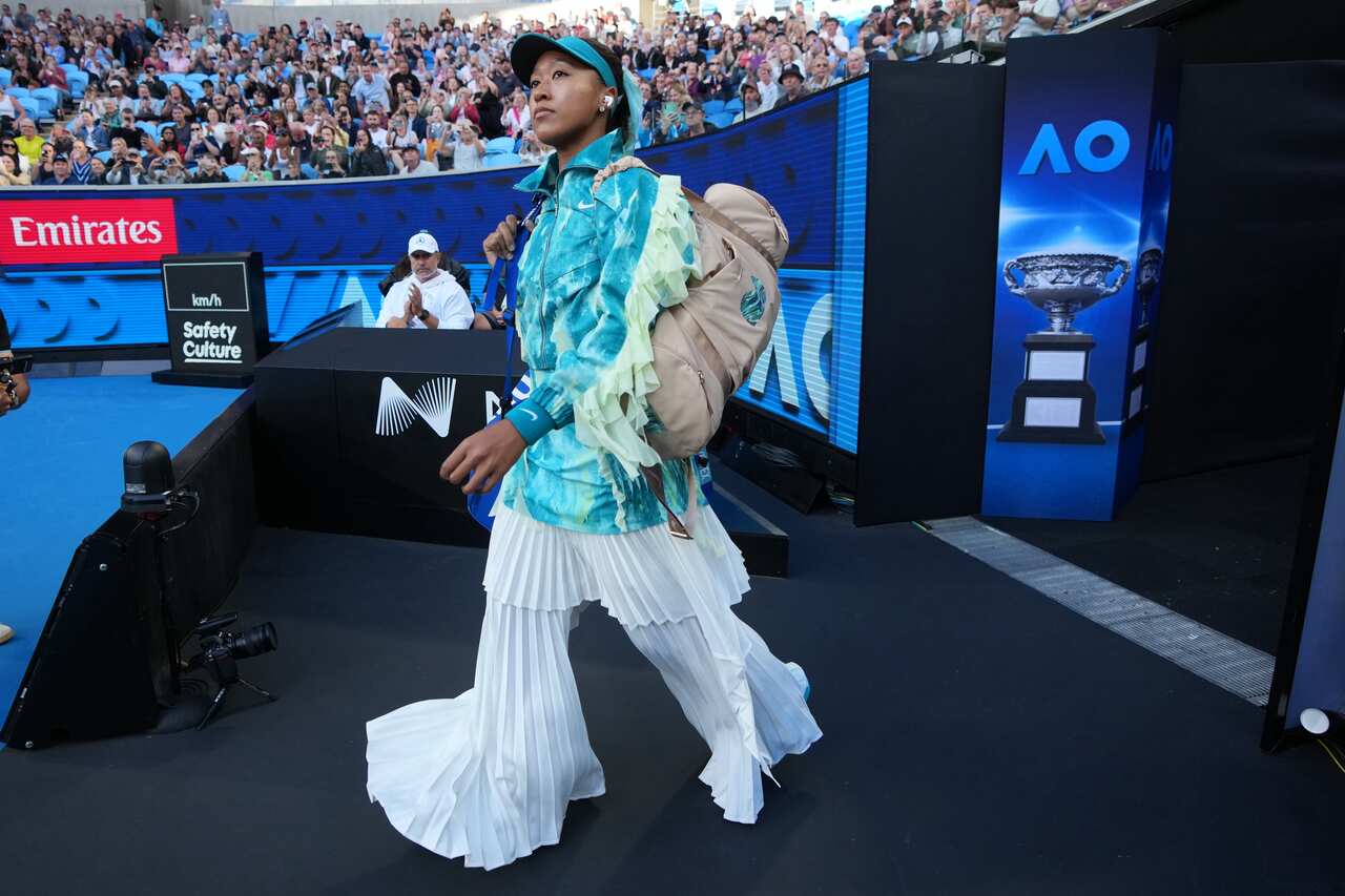 Tennis player Naomi Osaka walks onto a blue court at the Australian Open wearing a teal tie-dye jacket and a tiered, flowing white skirt while carrying a tan backpack.