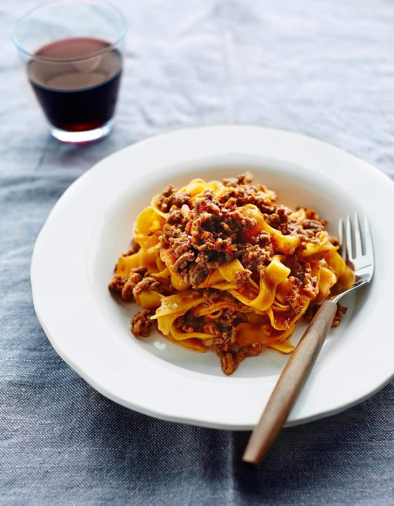 A white bowl with a wide edge sits on a blue tablecloth. It holds a mound of pasta and ragu. A fork sits on the side of the bowl.