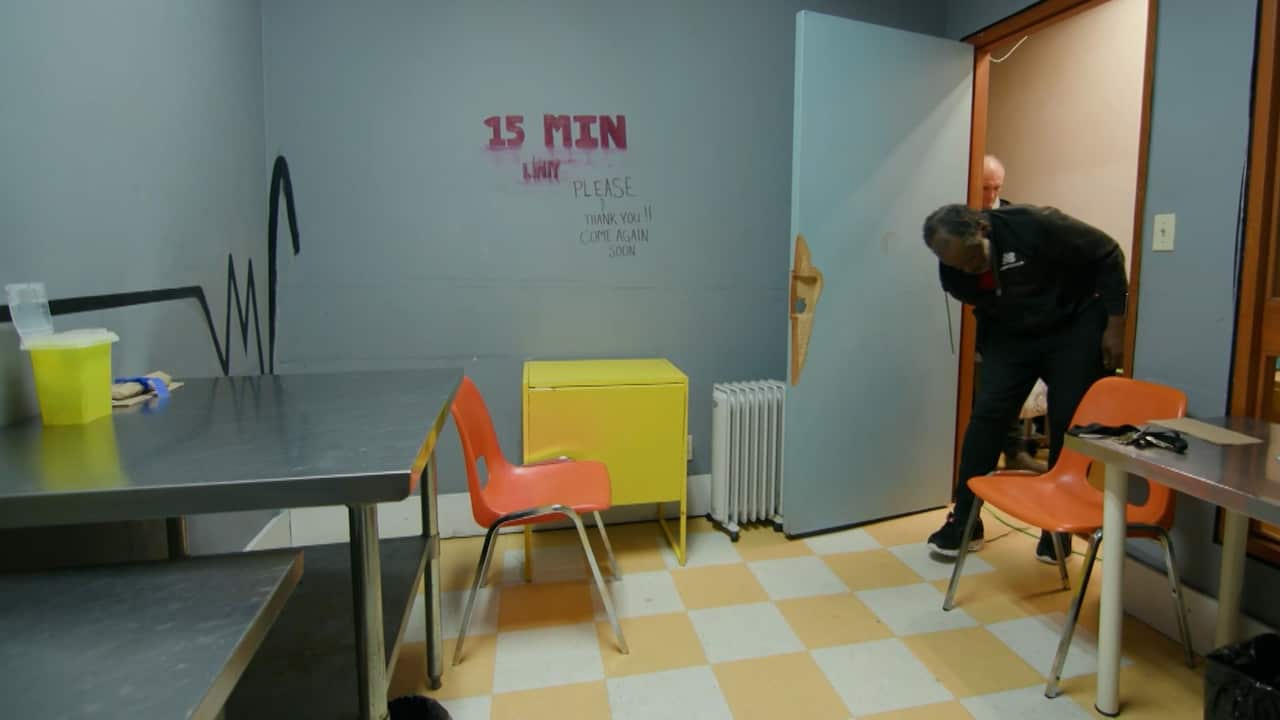 A man walks into a room for supervised drug consumption with a metal table, two orance chairs and a yellow cabinet inside 