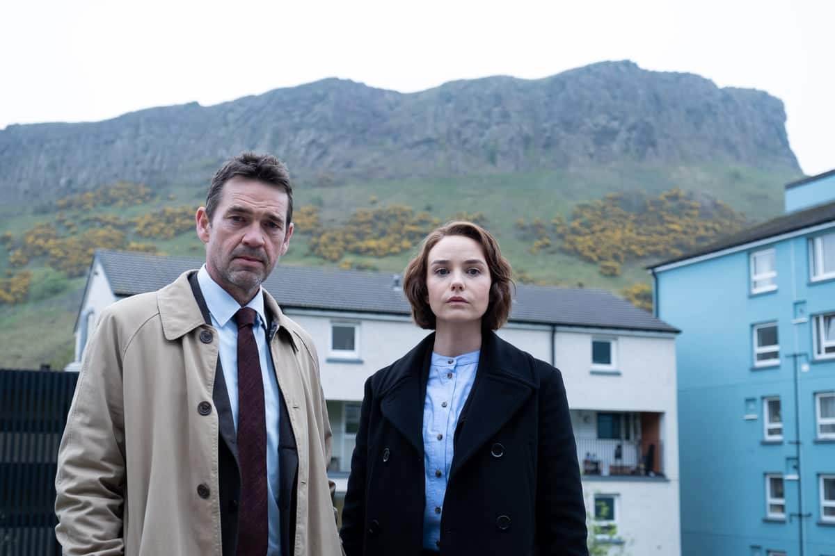 A man and a woman, both detectives stand in front of houses and mountains looking at the camera