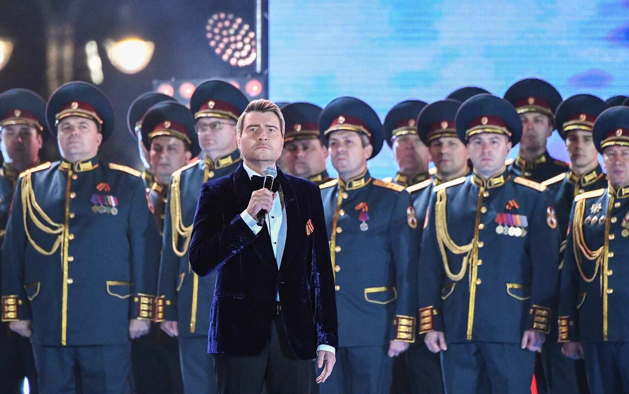 A man in a purple velvet jacket holds a microphone in front of men in military dress