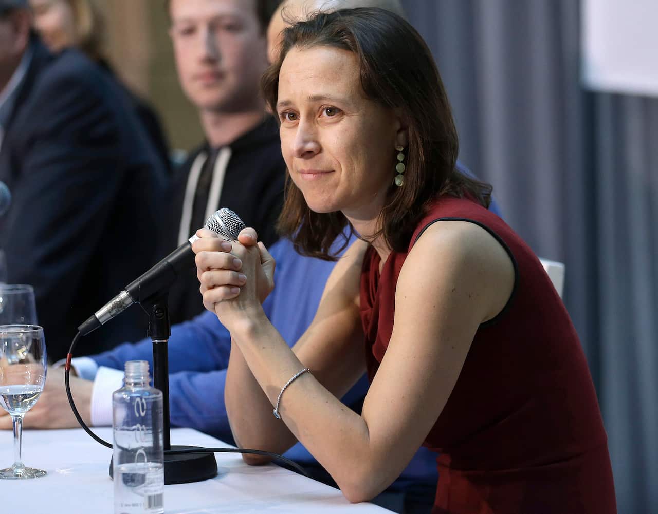 A woman with shoulder length brown hair wearing a maroon dress sitting at a table in front of a microphone.