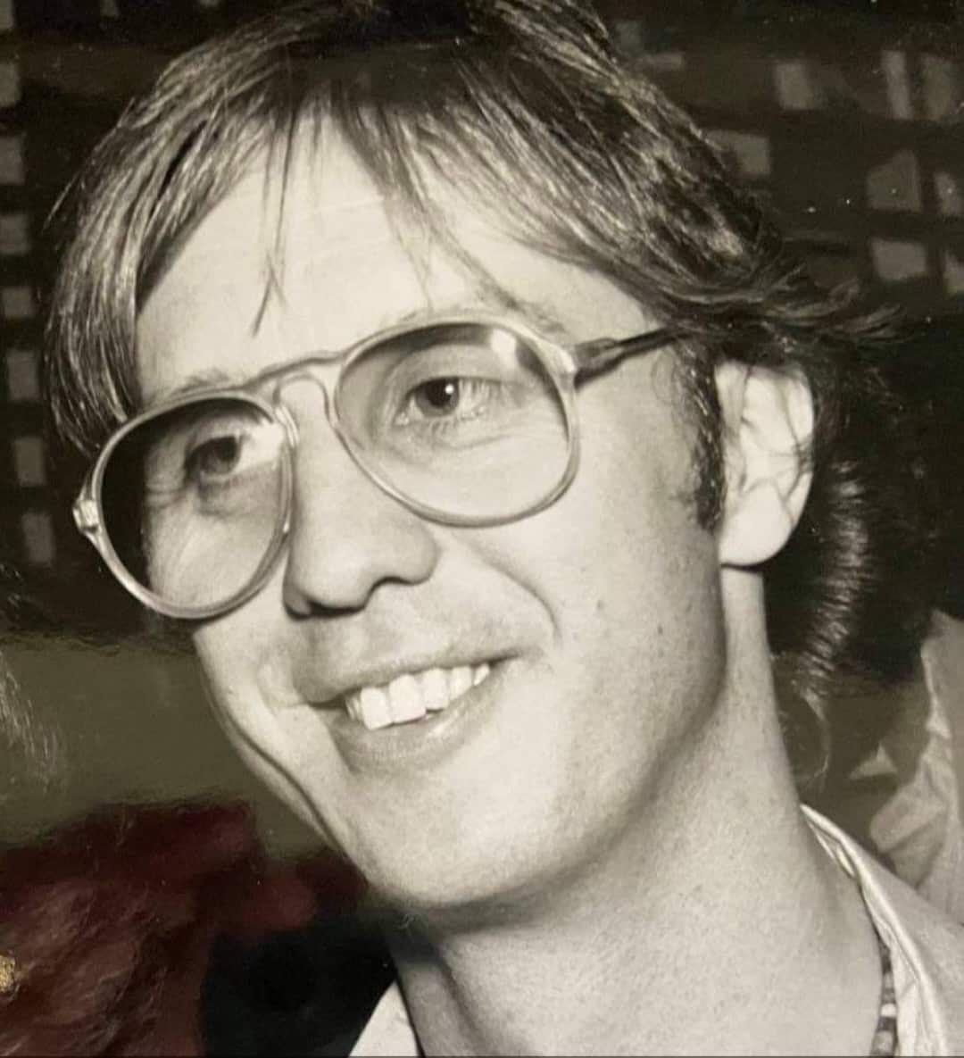 A black and white close-up portrait of a middle aged man wearing glasses and smiling.
