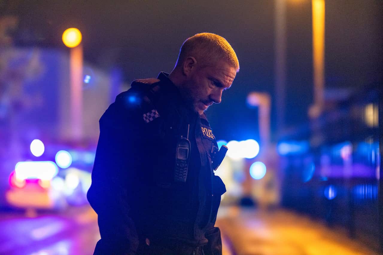 A police officer stands in the road at night, bright blue and red lights behind him