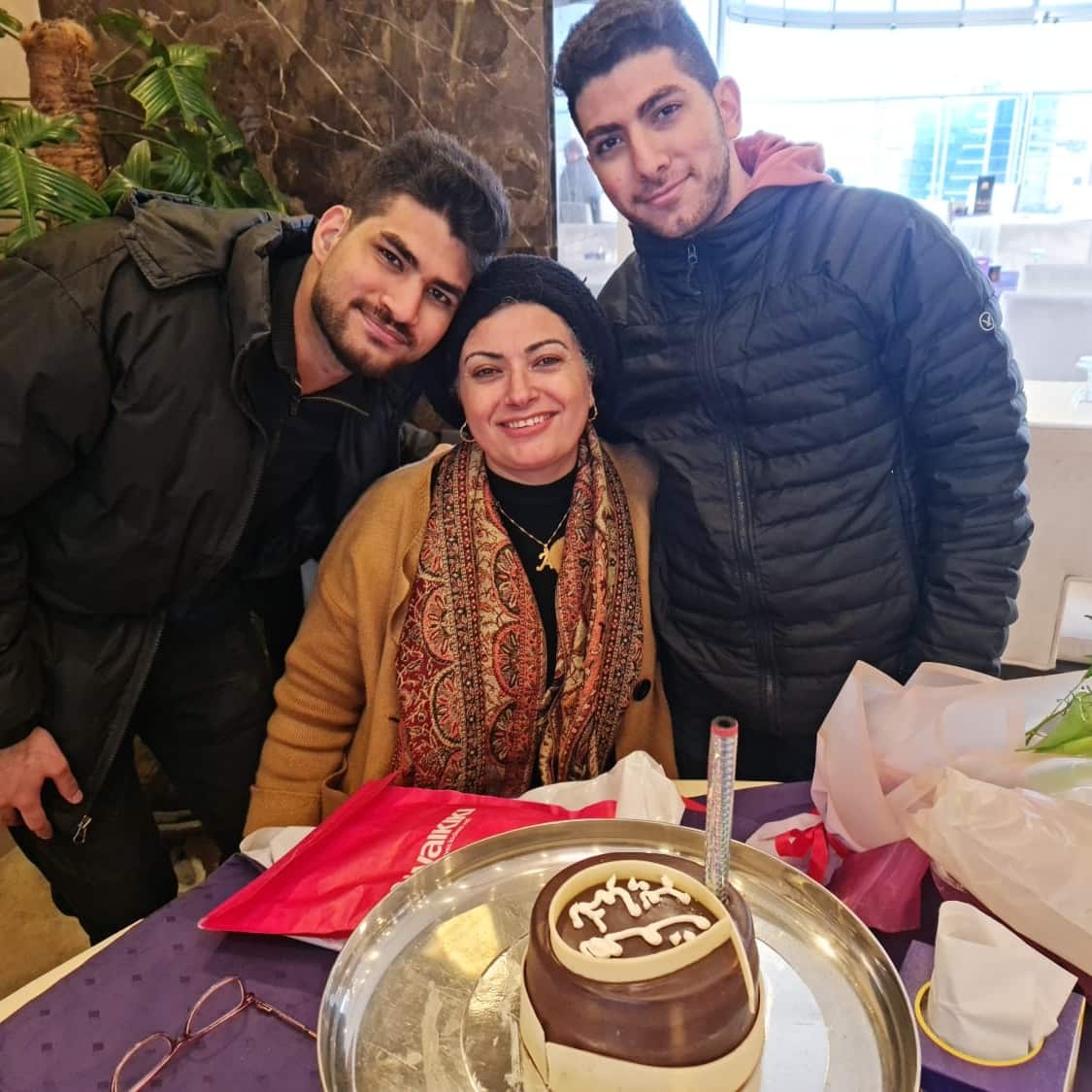 A woman with a headscarf sitting at a table with a silver tray and a cake on it, while two young men in black jackets stand by her sides hugging her