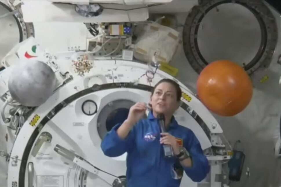 Nicole mann floating in her blue nasa uniform amongst the utilitarian cables and exposed metal of the ISS with her dream catcher floating before her