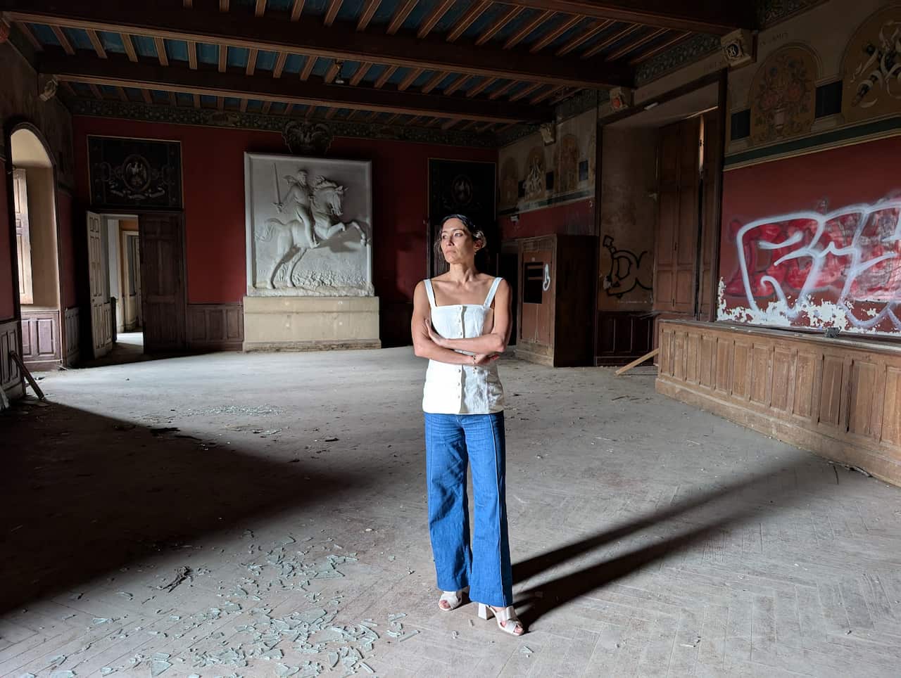 SBS presenter Kumi Taguchi stands in the middle of a large, mostly empty room. The room would have once been grand, with wood paneling, red wallpaper and a large statue of a man riding a horse against the far wall, but the floor is covered in glass and there is graffiti on the wall.