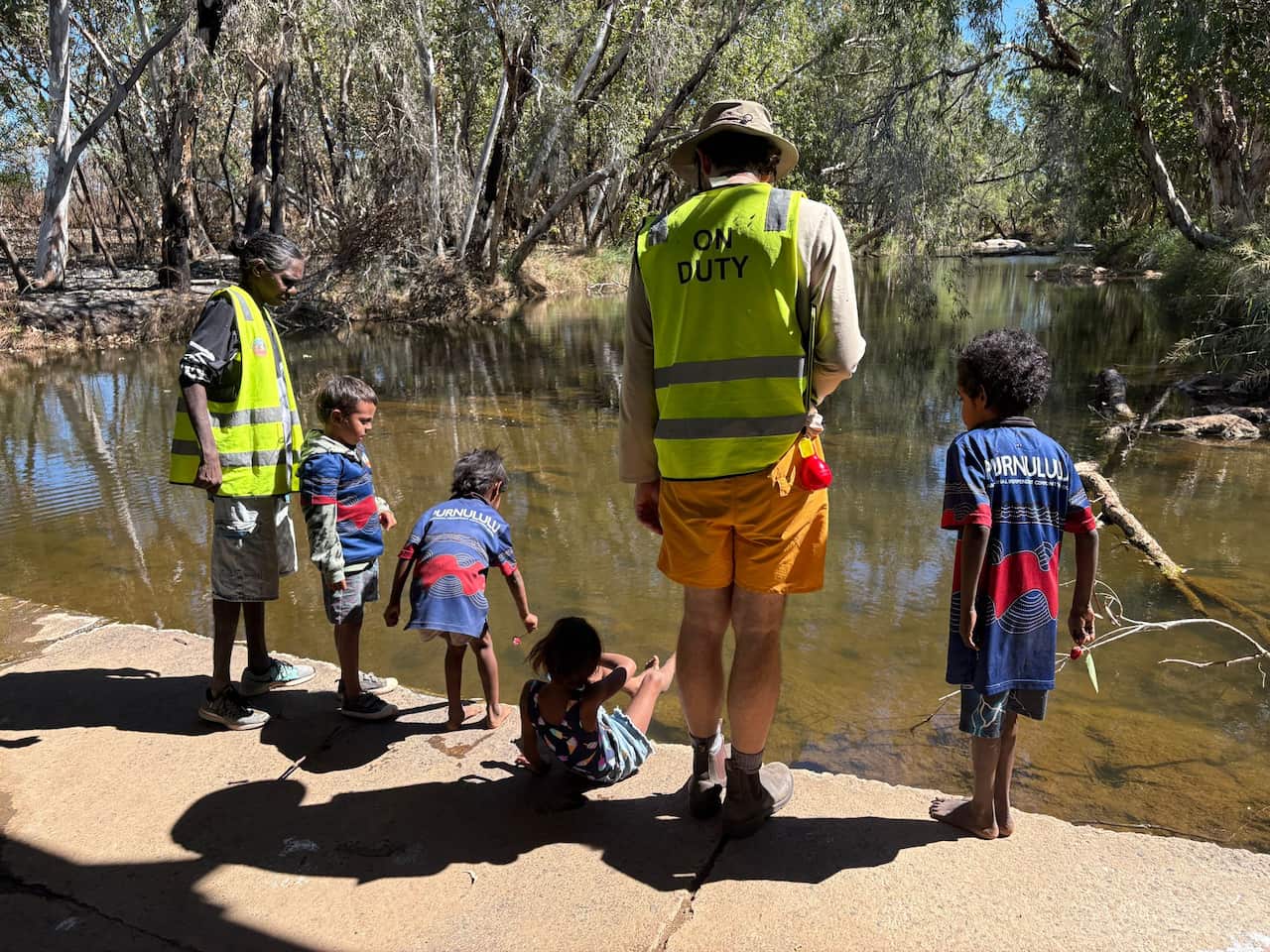 Two teachers wearing bright yellow safety vests stand with four young children wearing blue and red polo shirts on a concrete path next to a creek