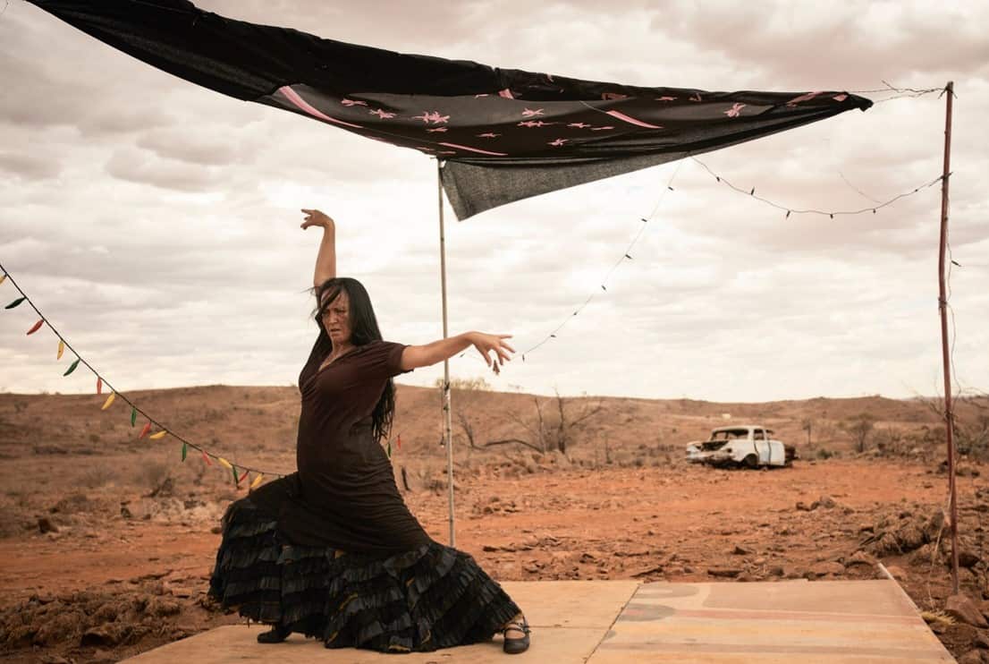 Marina Tamayo wearing a black dress and dacing on a makeshift stage on a barren landscape