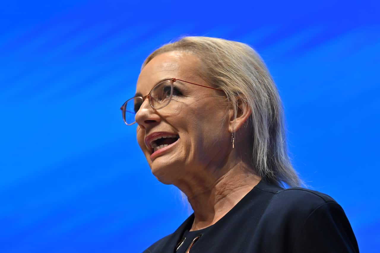 A woman with blonde hair speaking in front of a blue background.