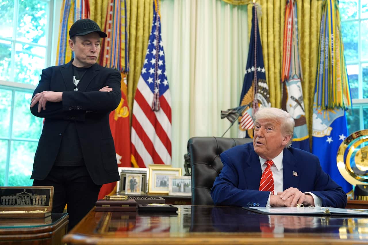 Donald Trump seated in the Oval Office. Elon Musk is standing near him.